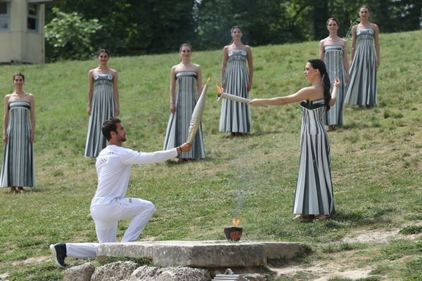 扮演最高女祭司的演員瑪麗·米娜（前右）點(diǎn)燃第一棒火炬手——希臘運(yùn)動(dòng)員、東京奧運(yùn)會(huì)男子賽艇項(xiàng)目金牌得主斯特凡諾斯·恩圖斯科斯手中的火炬。新華社記者 趙丁喆 攝